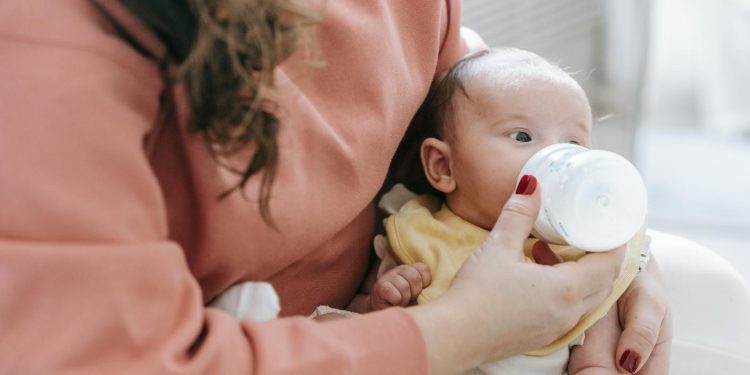 Crop anonymous mother in casual clothes feeding adorable newborn baby with bottle while sitting on sofa in light room at home